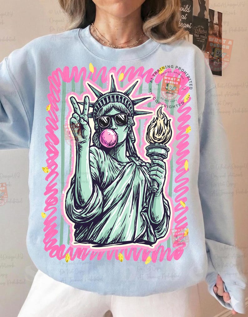 Puede incluir: Sudadera azul claro con un gr&aacute;fico de la Estatua de la Libertad con gafas de sol y soplando una burbuja. La estatua sostiene una antorcha y hace el signo de la paz. El dise&ntilde;o est&aacute; enmarcado en rosa con detalles amarillos.