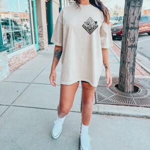 May include: A woman wearing a beige oversized t-shirt with a black graphic of a palm tree and waves in a diamond shape. She is wearing white trainers and has a tattoo on her leg.