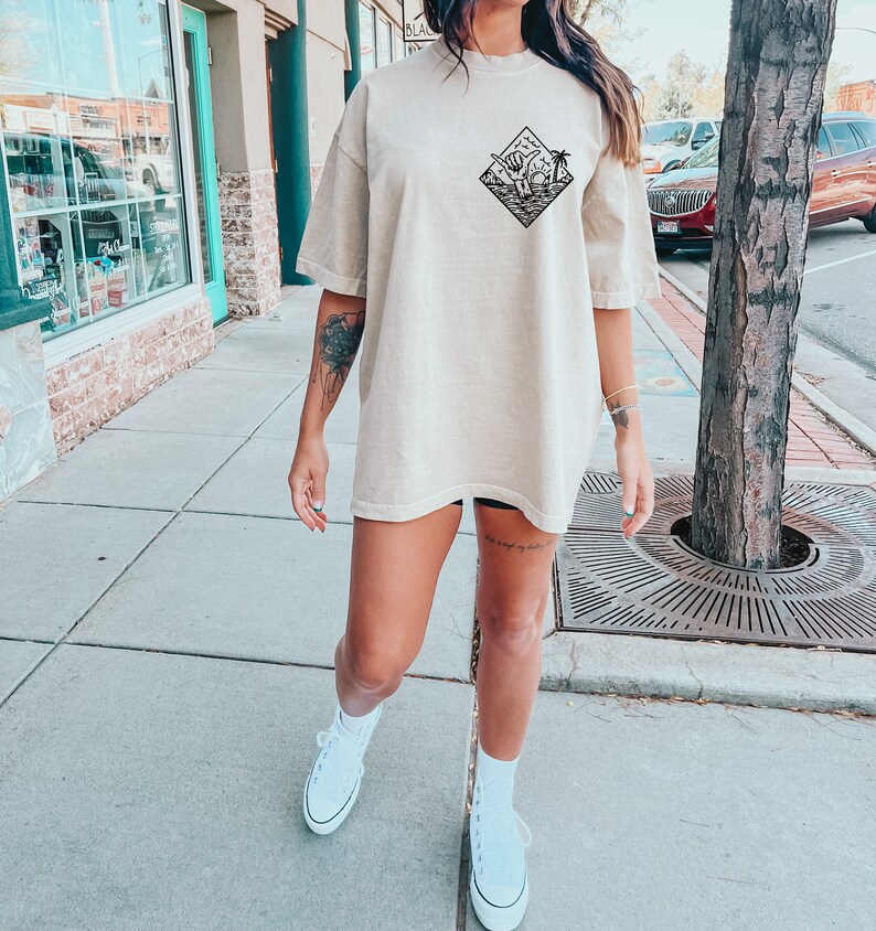 May include: A woman wearing a beige oversized t-shirt with a black graphic of a palm tree and waves in a diamond shape. She is wearing white trainers and has a tattoo on her leg.