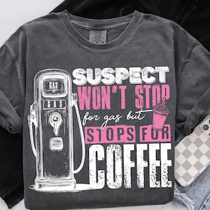 Op de afbeelding: Een grijs T-shirt met een vintage benzinepomp afbeelding en de tekst "SUSPECT WON'T STOP for gas but STOPS FOR COFFEE" in roze en wit.