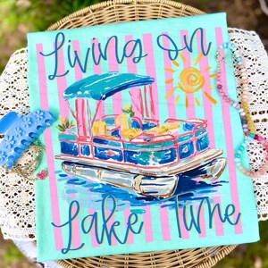 May include: A turquoise and pink striped canvas features a pontoon boat illustration with the words "Living on Lake Time" in cursive. The boat has a blue canopy and is surrounded by water and a yellow sun.