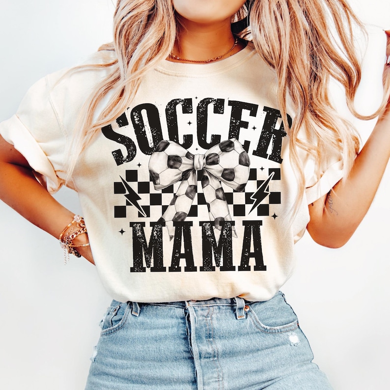 Puede incluir: Una camiseta blanca con el texto "SOCCER MAMA" en negro con un patr&oacute;n de cuadros blanco y negro y un lazo de bal&oacute;n de f&uacute;tbol blanco y negro.