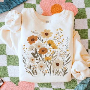 May include: A cream-colored sweatshirt with a floral print featuring yellow, orange, and brown flowers. The sweatshirt has a crew neck and long sleeves. The text "Comfort Colors" is printed on the inside of the neck.