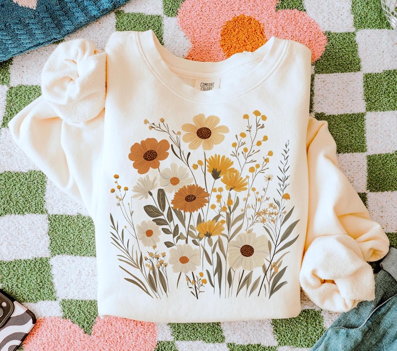 May include: A cream-colored sweatshirt with a floral print featuring yellow, orange, and brown flowers. The sweatshirt has a crew neck and long sleeves. The text "Comfort Colors" is printed on the inside of the neck.
