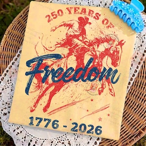 May include: A tan t-shirt with a red and blue graphic of a cowboy riding a horse, with the word "Freedom" in blue script. The text "250 Years of" and "1776-2026" are also visible. A blue hair clip with flower details is in the upper right corner.