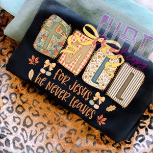 May include: Black t-shirt with the words "FAM for Jesus he Never Leaves" in orange script. The letters are designed as gifts with floral, plaid, and patterned backgrounds, tied with yellow bows. Autumn leaves and pumpkins decorate the design.