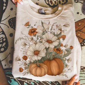 Pu&ograve; includere: Felpa color crema con un design floreale con fiori bianchi e arancioni e due zucche. Il design &egrave; stampato sulla parte anteriore della felpa piegata. La felpa &egrave; tenuta da una persona.