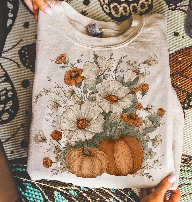 Pu&ograve; includere: Felpa color crema con un design floreale con fiori bianchi e arancioni e due zucche. Il design &egrave; stampato sulla parte anteriore della felpa piegata. La felpa &egrave; tenuta da una persona.