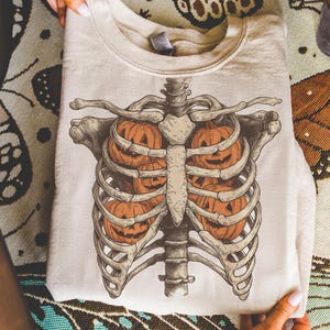 May include: A cream-colored sweatshirt featuring a detailed illustration of a rib cage with jack-o'-lanterns inside. The design is in shades of orange, brown, and beige, perfect for Halloween or fall. The sweatshirt is folded and held by hands.