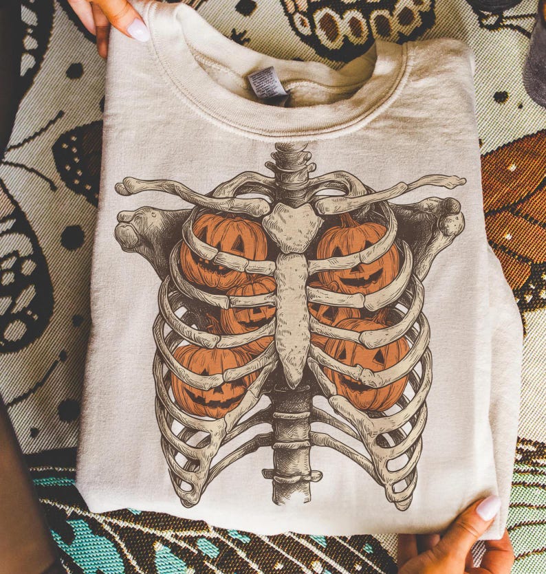May include: A cream-colored sweatshirt featuring a detailed illustration of a rib cage with jack-o'-lanterns inside. The design is in shades of orange, brown, and beige, perfect for Halloween or fall. The sweatshirt is folded and held by hands.