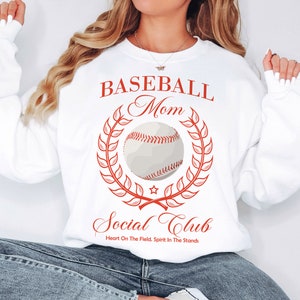 Peut inclure: Sweat-shirt blanc avec un texte rouge qui dit "Baseball Mom Social Club" avec un graphique de baseball et une couronne de laurier. Le texte sous le graphique dit "Heart On The Field. Spirit In The Stands."