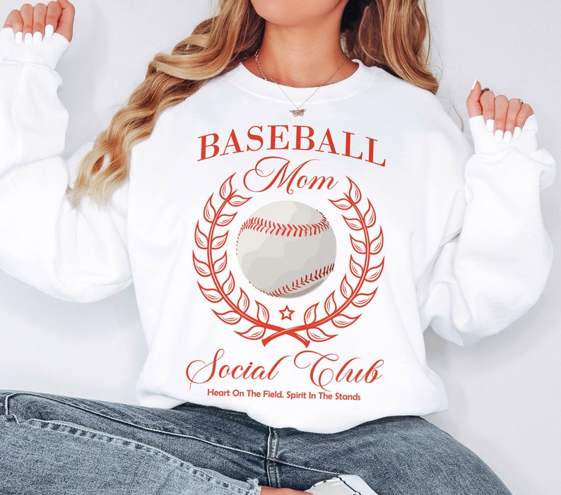 Peut inclure: Sweat-shirt blanc avec un texte rouge qui dit "Baseball Mom Social Club" avec un graphique de baseball et une couronne de laurier. Le texte sous le graphique dit "Heart On The Field. Spirit In The Stands."