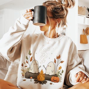 May include: Cream-colored sweatshirt featuring a whimsical illustration of two ghost figures roasting marshmallows over a campfire. The design includes a pumpkin, mushrooms, and fall foliage. The person in the image is holding a black mug.