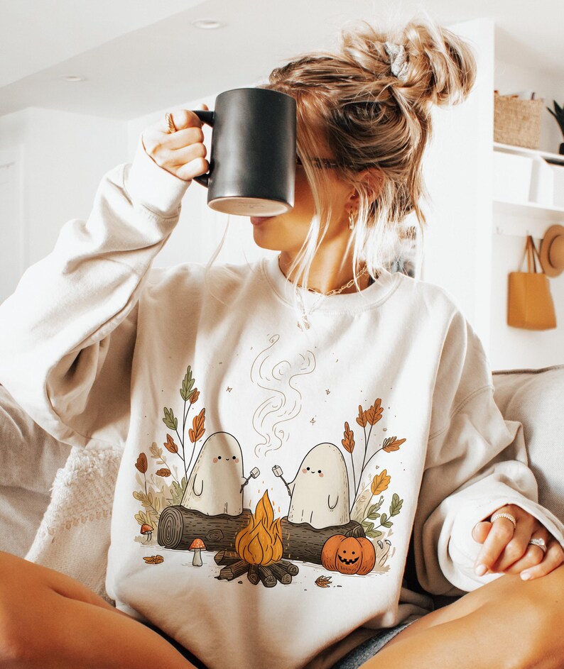 May include: Cream-colored sweatshirt featuring a whimsical illustration of two ghost figures roasting marshmallows over a campfire. The design includes a pumpkin, mushrooms, and fall foliage. The person in the image is holding a black mug.