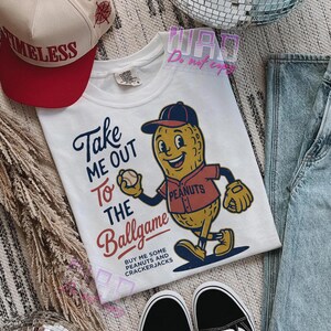 May include: White t-shirt with a baseball-themed graphic. The design features a cartoon peanut character in a baseball uniform holding a baseball, with the text "Take Me Out To The Ballgame". A red and white hat is visible.
