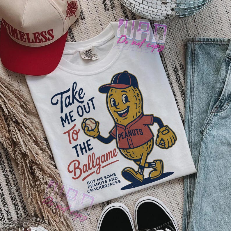 May include: White t-shirt with a baseball-themed graphic. The design features a cartoon peanut character in a baseball uniform holding a baseball, with the text "Take Me Out To The Ballgame". A red and white hat is visible.