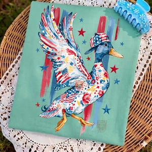 May include: A decorative cushion featuring a duck design with American flag motifs. The duck is depicted wearing a cap and has wings decorated with stars and stripes. The background is a pale teal with red and blue stripes and stars.