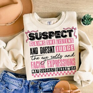 May include: A white sweatshirt with a black and pink graphic print that says "SUSPECT CLAIMS SHE LISTENS AND DOESNT JUDGE the eye rolls and FACIAL EXPRESSIONS MAY SUGGEST OTHERWISE".
