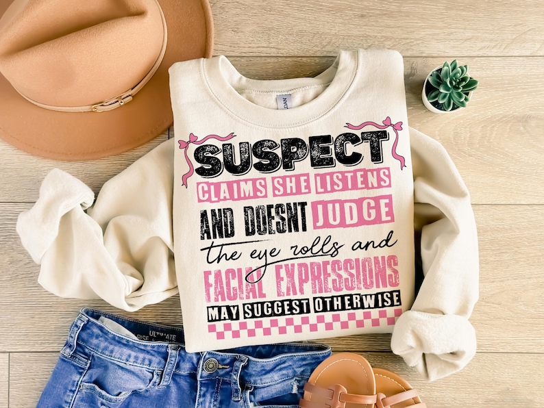 May include: A white sweatshirt with a black and pink graphic print that says "SUSPECT CLAIMS SHE LISTENS AND DOESNT JUDGE the eye rolls and FACIAL EXPRESSIONS MAY SUGGEST OTHERWISE".
