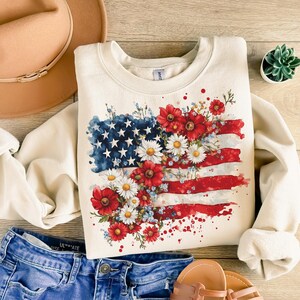 May include: A cream-colored sweatshirt with a watercolor design of an American flag with red, white, and blue flowers.