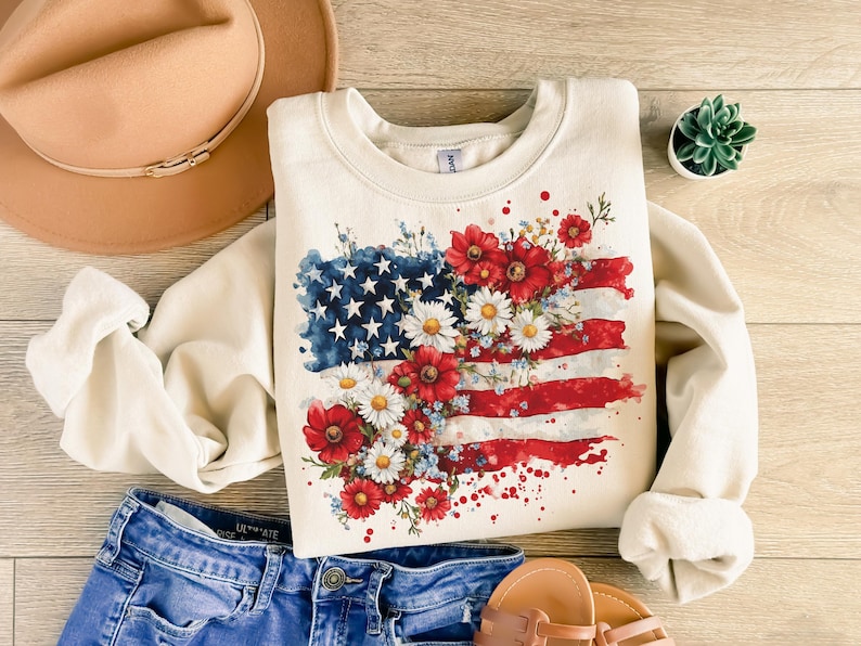 May include: A cream-colored sweatshirt with a watercolor design of an American flag with red, white, and blue flowers.