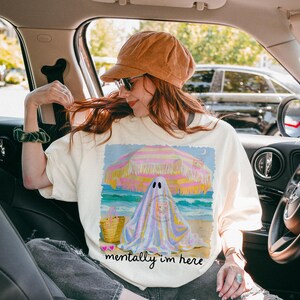 May include: An off-white t-shirt featuring a graphic of a ghost sitting under a yellow and pink striped beach umbrella on a sandy beach. The design includes a straw tote bag, a small crescent moon, and the text "mentally i'm here" in black script.