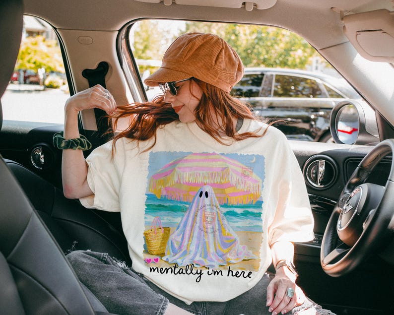 May include: An off-white t-shirt featuring a graphic of a ghost sitting under a yellow and pink striped beach umbrella on a sandy beach. The design includes a straw tote bag, a small crescent moon, and the text "mentally i'm here" in black script.