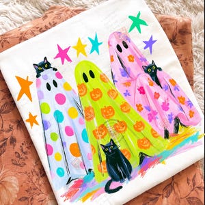 May include: A white t-shirt featuring a graphic print of three ghosts in colorful sheets. One ghost has polka dots, one has orange pumpkins, and one has floral patterns. Three black cats are peeking out from the sheets, surrounded by colorful stars.