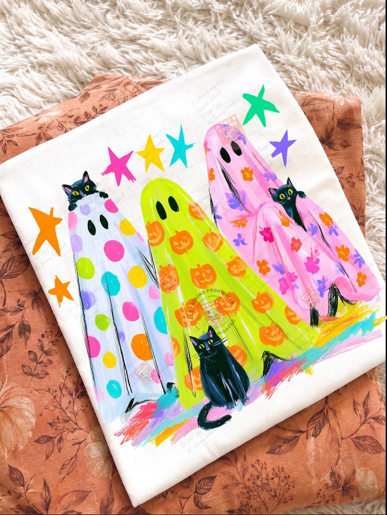 May include: A white t-shirt featuring a graphic print of three ghosts in colorful sheets. One ghost has polka dots, one has orange pumpkins, and one has floral patterns. Three black cats are peeking out from the sheets, surrounded by colorful stars.
