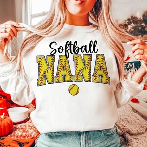 May include: White sweatshirt with the text "Softball Nana" in black and yellow letters with a red outline. The letters are decorated with a softball pattern. A yellow softball is below the text.