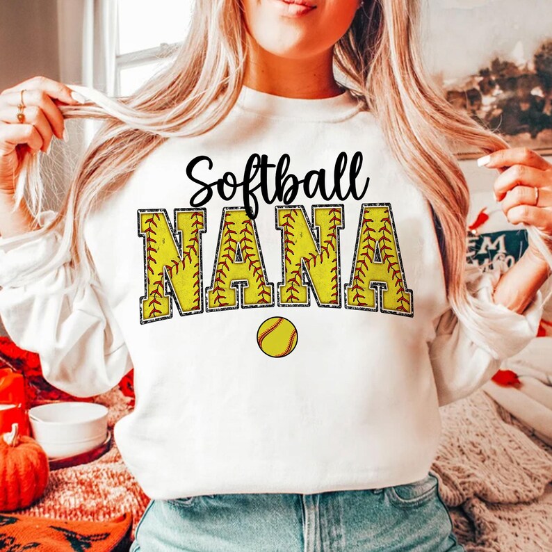 May include: White sweatshirt with the text "Softball Nana" in black and yellow letters with a red outline. The letters are decorated with a softball pattern. A yellow softball is below the text.