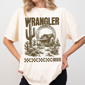 Pode incluir: Uma camiseta de cor creme com uma estampa gr&aacute;fica marrom de uma cena do deserto com uma casa, cactos e a palavra "WRANGLER" em letras grandes.