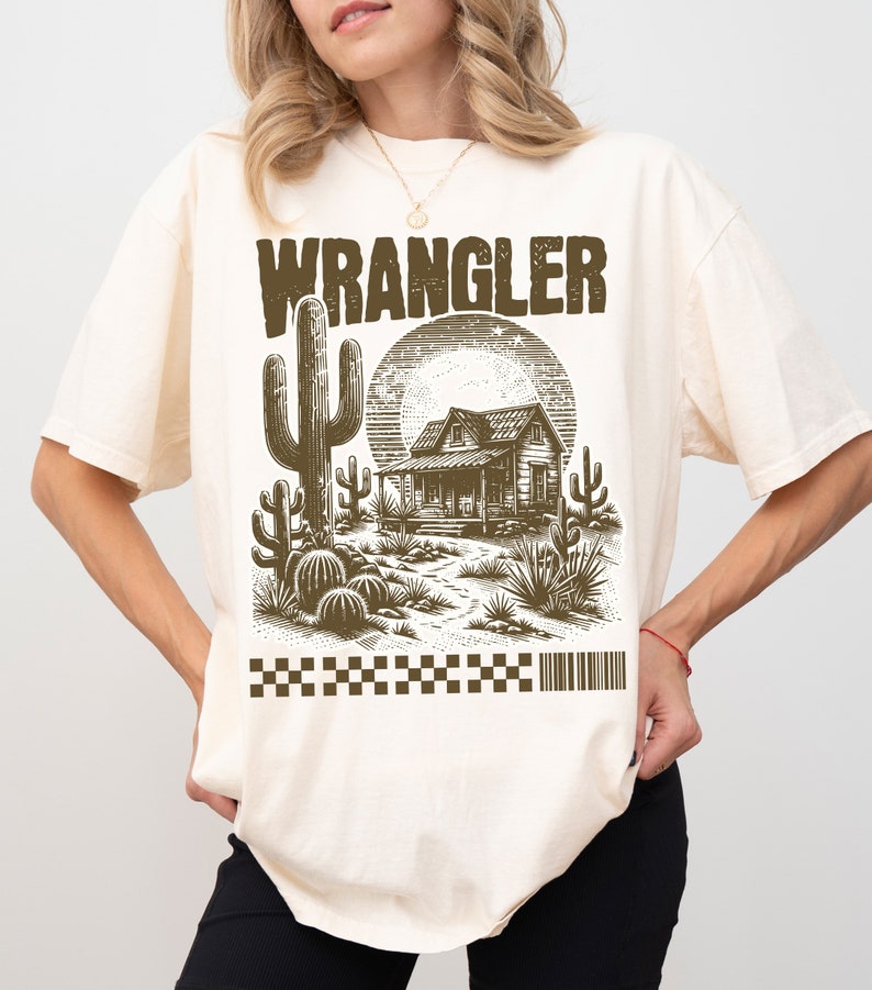 Pode incluir: Uma camiseta de cor creme com uma estampa gr&aacute;fica marrom de uma cena do deserto com uma casa, cactos e a palavra "WRANGLER" em letras grandes.