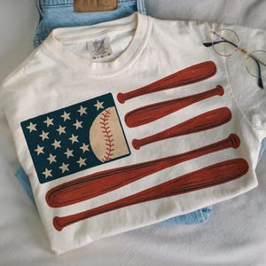 Peut inclure: T-shirt blanc cassé avec un motif de drapeau américain sur le thème du baseball. Les étoiles du drapeau sont blanches sur un rectangle bleu marine, et les rayures sont remplacées par des battes de baseball rouges. Une balle de baseball remplace les rayures du drapeau.