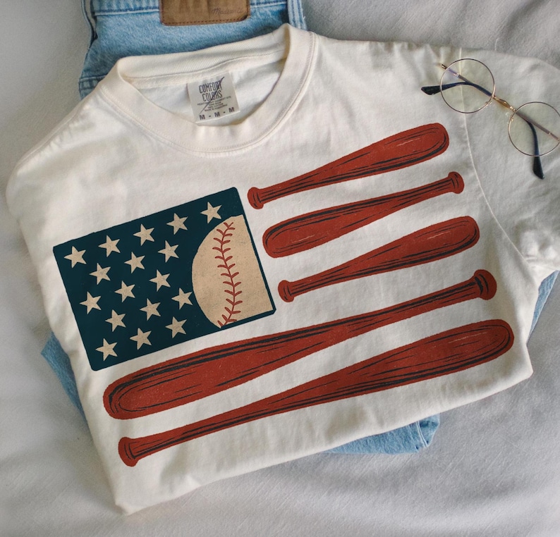 May include: Cream-colored t-shirt featuring a baseball-themed American flag design. The flag's stars are represented by white stars on a navy blue rectangle, and the stripes are replaced with red baseball bats. A baseball replaces the flag's stripes.