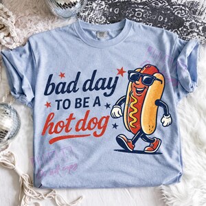 Puede incluir: Camiseta azul claro con la frase "bad day to be a hot dog" en azul marino y rojo. Un dibujo animado de un perrito caliente con gafas de sol caminando. La camiseta es de cuello redondo y manga corta.