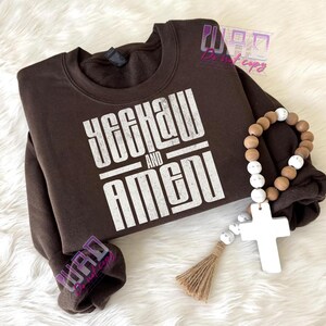 May include: A brown sweatshirt with the text "YEEHAW AND AMEN" in white block letters. A wooden bead rosary with a white cross and tassel is next to the sweatshirt.