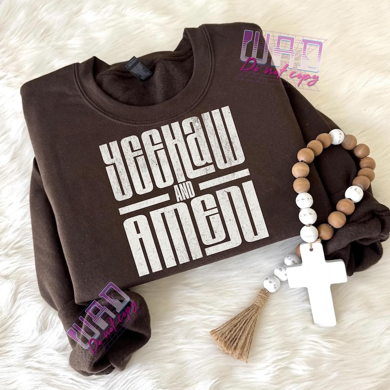May include: A brown sweatshirt with the text "YEEHAW AND AMEN" in white block letters. A wooden bead rosary with a white cross and tassel is next to the sweatshirt.