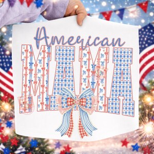 Op de afbeelding: Wit grafisch T-shirt met de tekst "American MAMA" in blauwe letters en blokletters. De letters zijn versierd met rode, witte en blauwe strikken en ruitpatronen. Onderaan een grote strik met rode, witte en blauwe accenten.