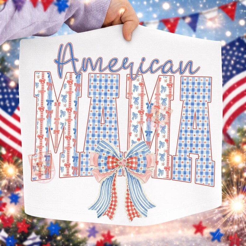 Op de afbeelding: Wit grafisch T-shirt met de tekst "American MAMA" in blauwe letters en blokletters. De letters zijn versierd met rode, witte en blauwe strikken en ruitpatronen. Onderaan een grote strik met rode, witte en blauwe accenten.