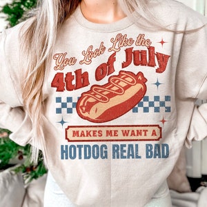 May include: A beige sweatshirt with a vintage-style graphic of a hot dog and the text "You Look Like the 4th of July Makes Me Want a Hotdog Real Bad".