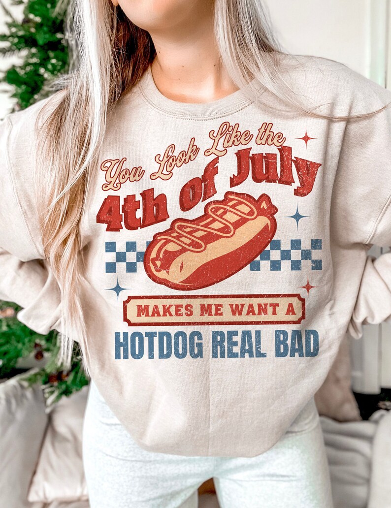 May include: A beige sweatshirt with a vintage-style graphic of a hot dog and the text "You Look Like the 4th of July Makes Me Want a Hotdog Real Bad".