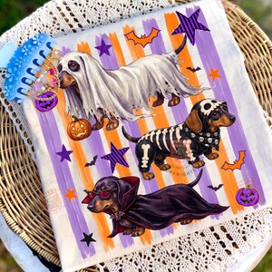 May include: A white fabric square featuring three dachshunds in Halloween costumes: one as a ghost with a pumpkin, one as a skeleton with a skull cap, and one as a vampire with red sunglasses. The background has orange and purple vertical stripes.