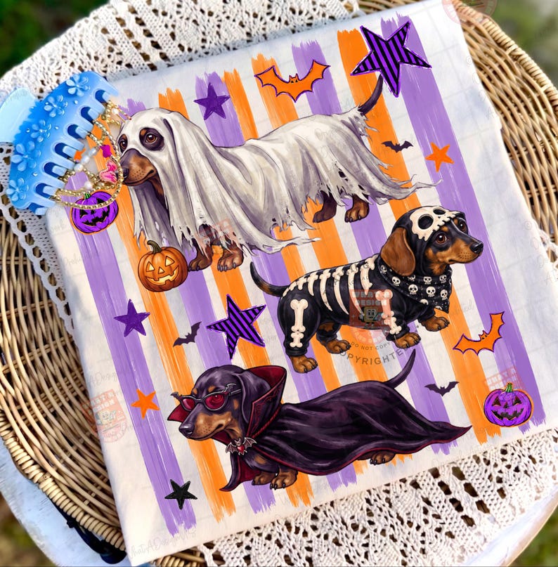 May include: A white fabric square featuring three dachshunds in Halloween costumes: one as a ghost with a pumpkin, one as a skeleton with a skull cap, and one as a vampire with red sunglasses. The background has orange and purple vertical stripes.