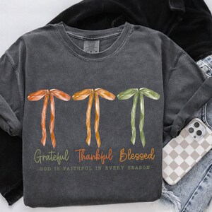 May include: Dark gray t-shirt with watercolor bows in orange, yellow, and green. The text reads "Grateful, Thankful, Blessed" with "God is faithful in every season" below. A phone with a checkered case and jeans are visible.