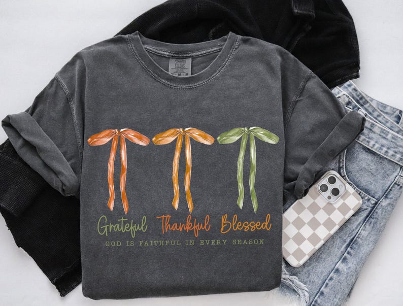 May include: Dark gray t-shirt with watercolor bows in orange, yellow, and green. The text reads "Grateful, Thankful, Blessed" with "God is faithful in every season" below. A phone with a checkered case and jeans are visible.