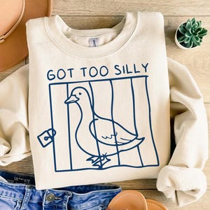 Op de afbeelding: Een crèmekleurig sweatshirt met een blauwe afbeelding van een vogel in een kooi. De tekst "GOT TOO SILLY" staat boven de afbeelding.