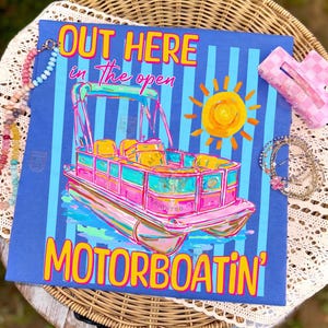 May include: A blue t-shirt with a colorful illustration of a pontoon boat, a sun, and the words "OUT HERE in the open MOTORBOATIN'". The boat is pink, teal, and yellow. A pink hair clip and beaded necklaces are in the image.
