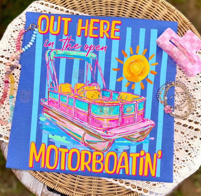 May include: A blue t-shirt with a colorful illustration of a pontoon boat, a sun, and the words "OUT HERE in the open MOTORBOATIN'". The boat is pink, teal, and yellow. A pink hair clip and beaded necklaces are in the image.