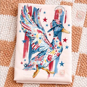 May include: A folded, pale pink fabric displays a whimsical duck illustration. The duck is decorated with patriotic patterns, including the American flag, stars, and stripes. It wears a matching cap and has red, white, and blue accents.